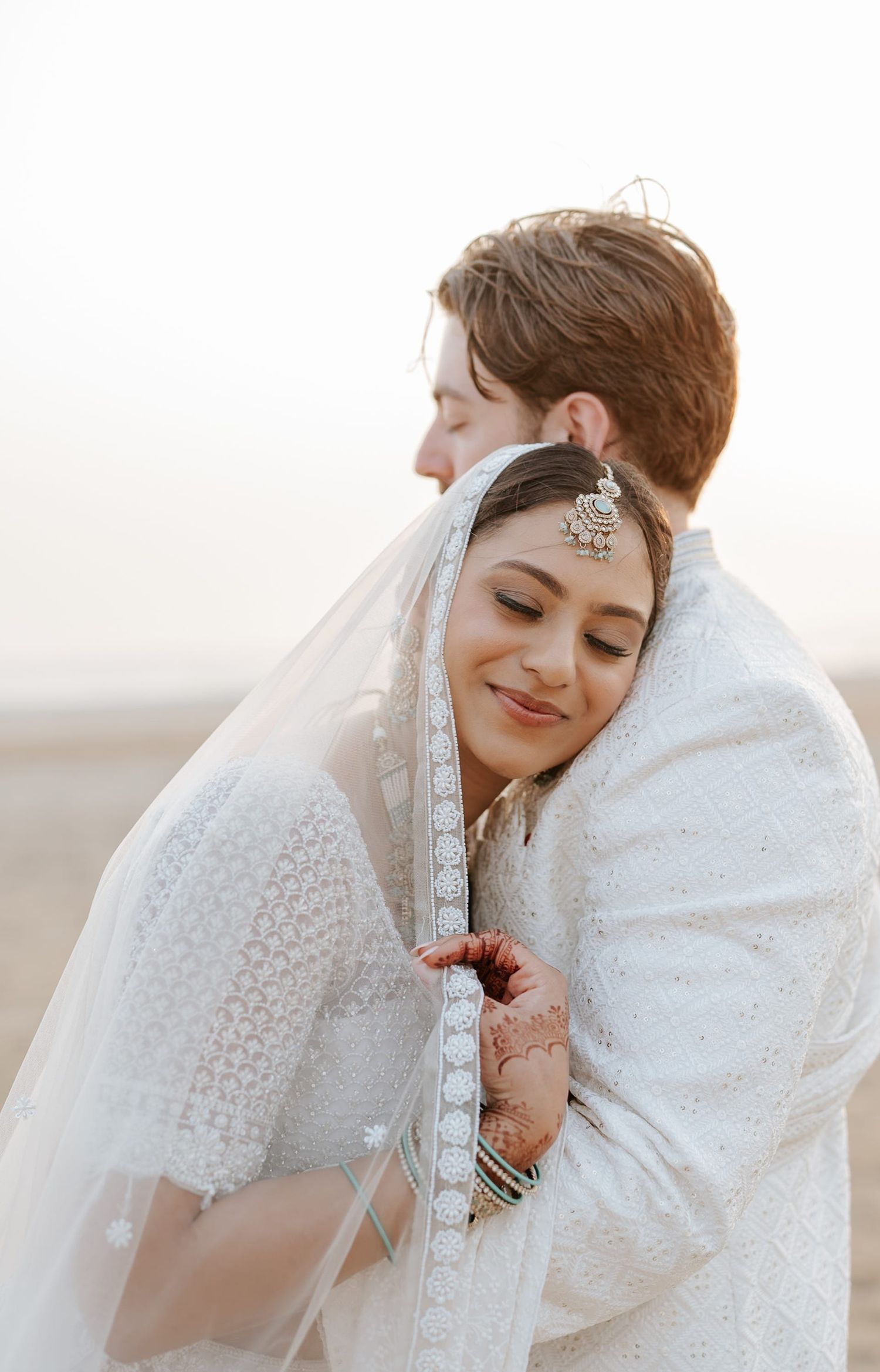Bride in white embroidered saree with mehndi and silver jewelry embraced by groom in soft natural light.