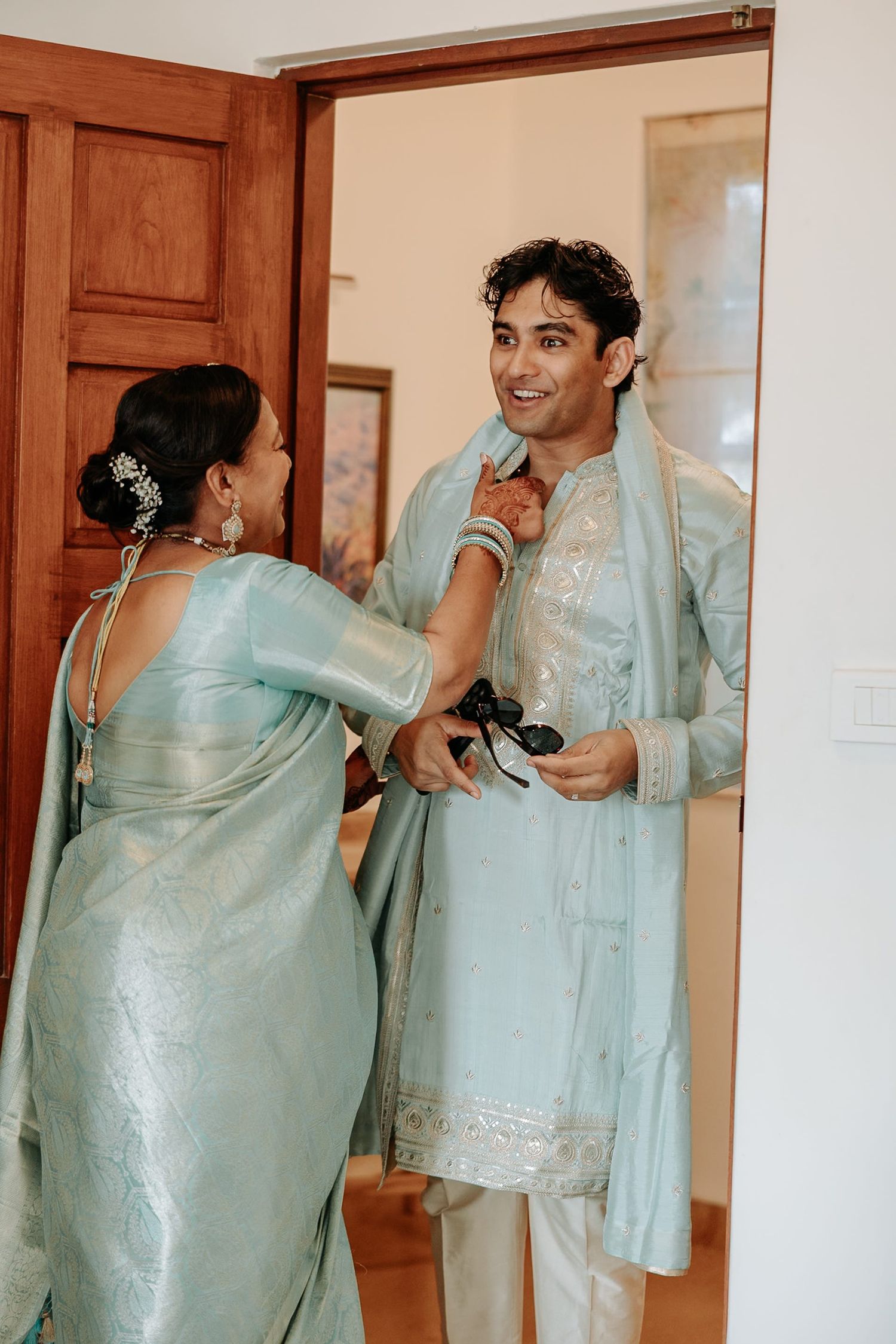 Indian groom in light blue kurta being helped by woman in matching saree during wedding preparation.