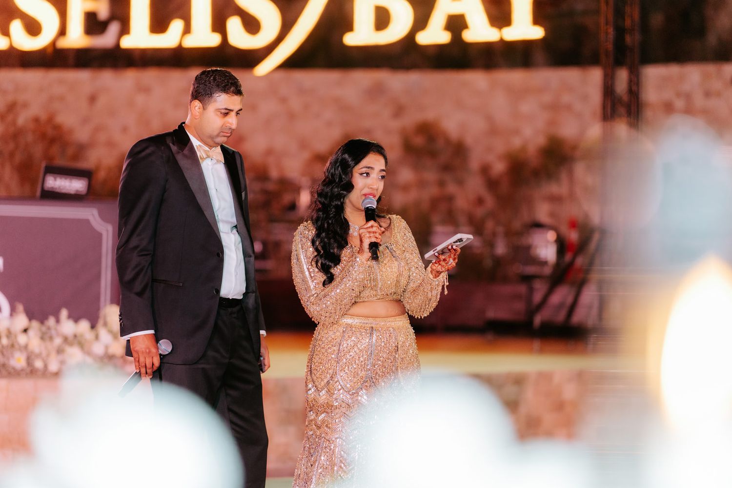 A woman in a gold sequin gown speaks into a microphone on stage beside a man in a tuxedo at an elegant event.
