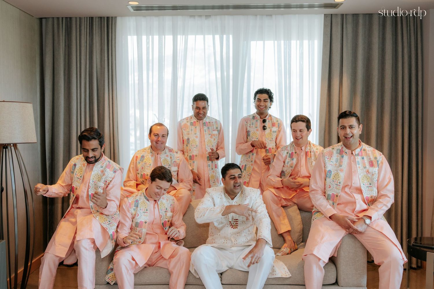 Groom in white kurta poses with groomsmen in matching pink floral sherwanis in a bright hotel room.