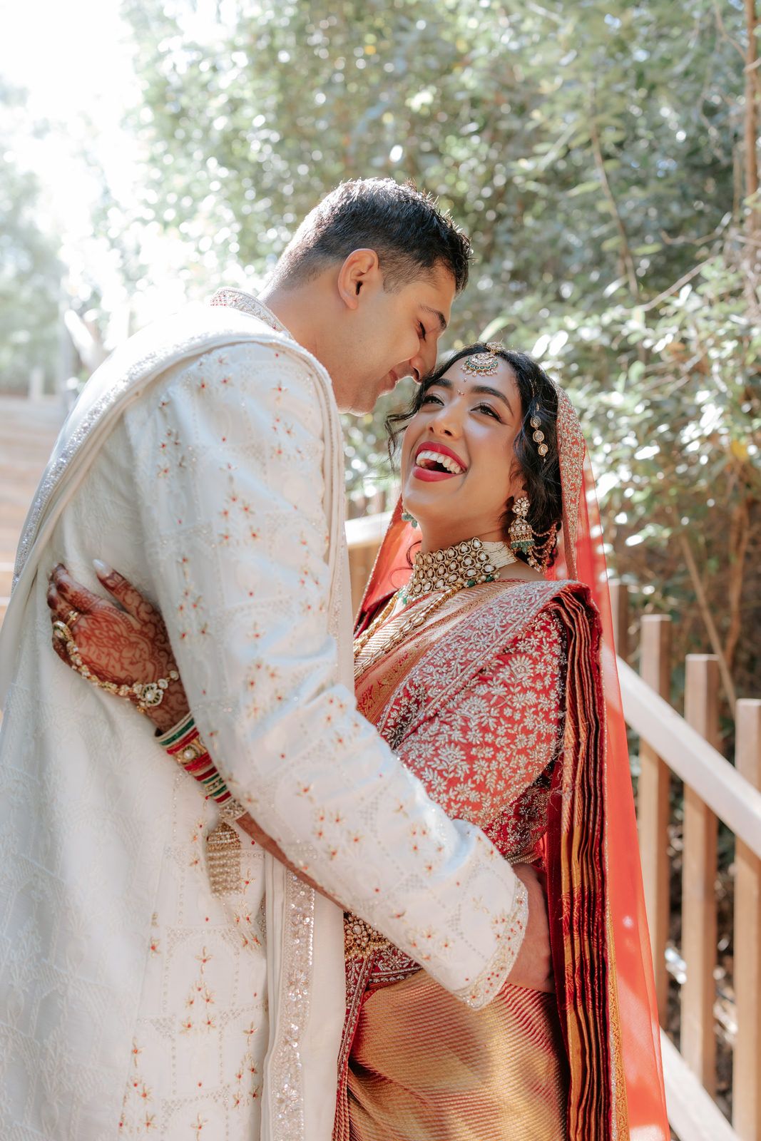 Joyful Indian couple embracing outdoors, bride in red saree and groom in white sherwani, laughing.