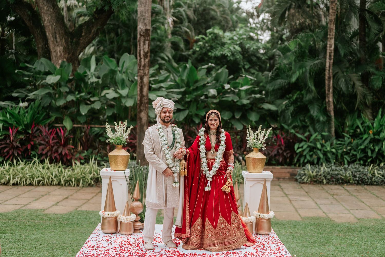 Indian bride and groom in traditional red and gold wedding attire stand together outdoors surrounded by lush tropical greenery.