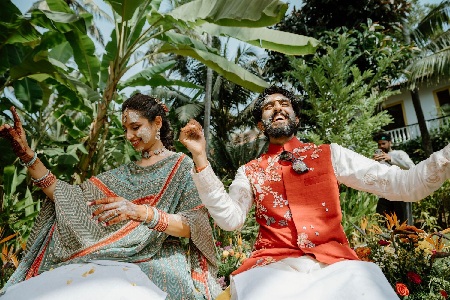 Indian couple in traditional attire dancing joyfully outdoors, surrounded by lush tropical greenery.