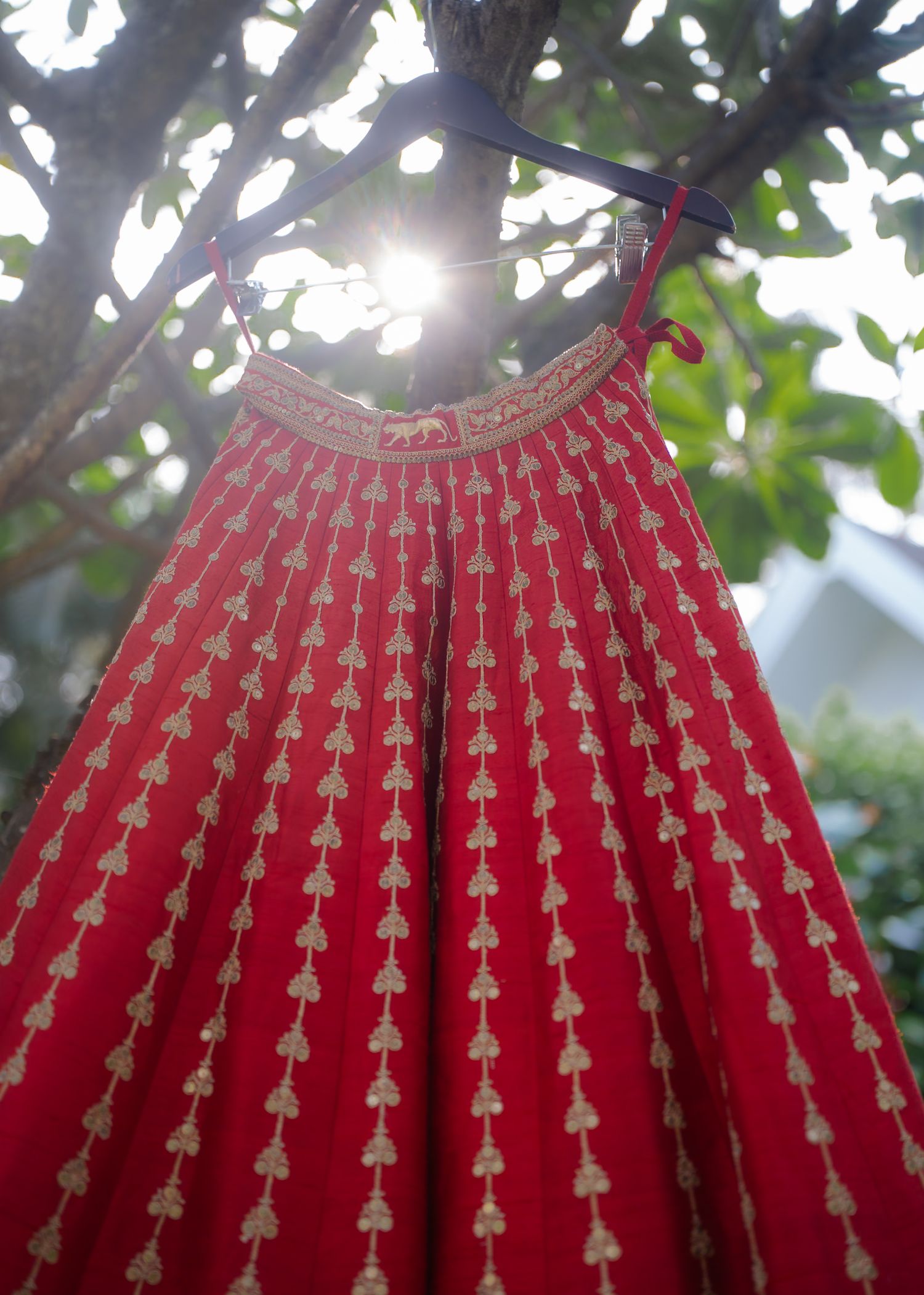 Red traditional Indian lehenga with gold embroidery hanging on a hanger outdoors among green trees.