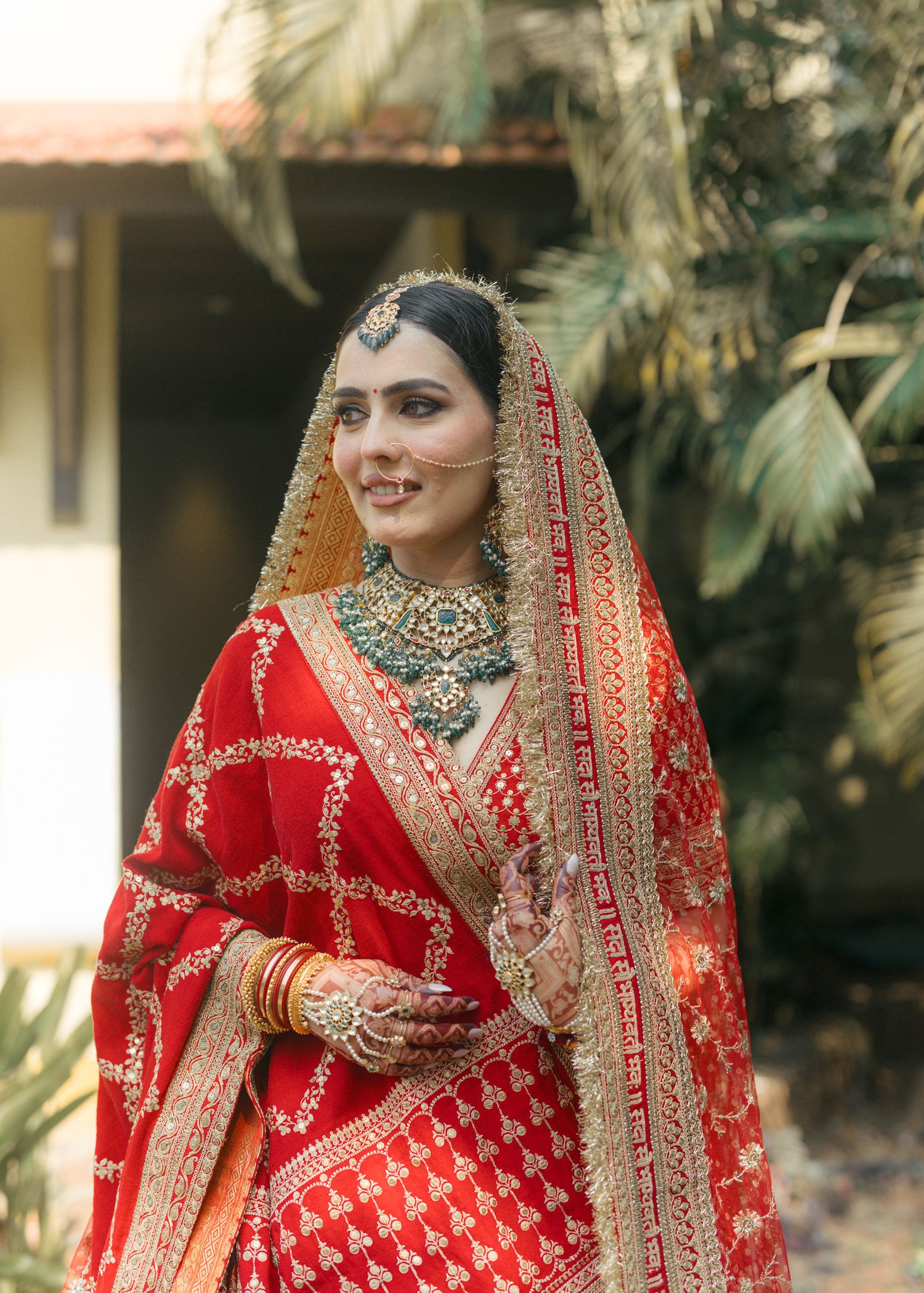 A bride in a red and gold embroidered lehenga with green kundan jewelry and dupatta smiles outdoors.