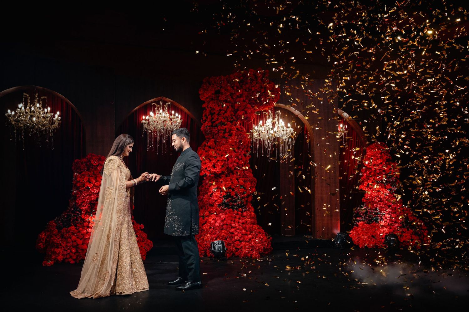 Couple holding hands on stage surrounded by red floral arches and golden confetti at an elegant wedding celebration.
