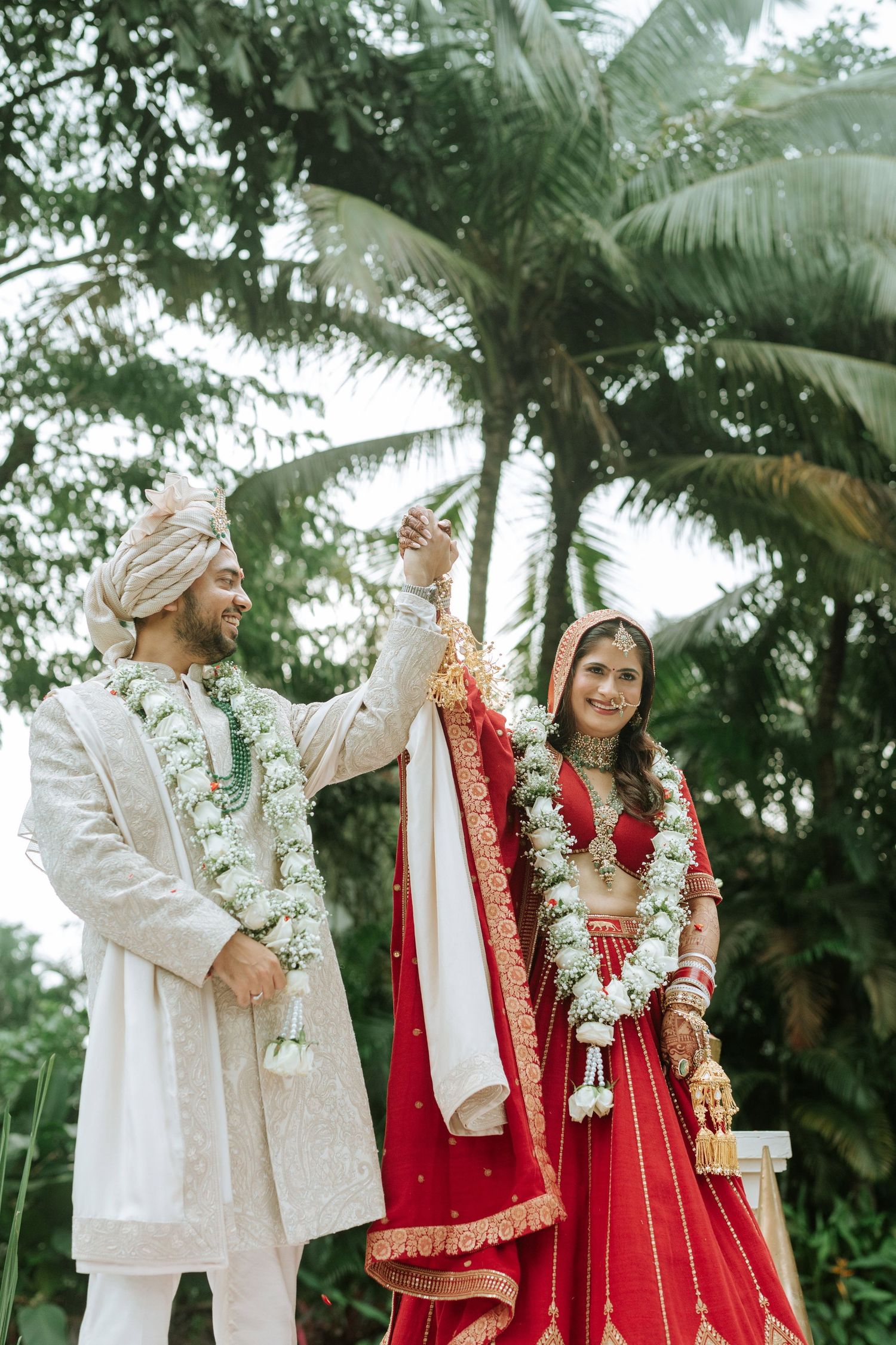 Indian couple in traditional wedding attire, groom in white sherwani and bride in red lehenga, celebrating outdoors.