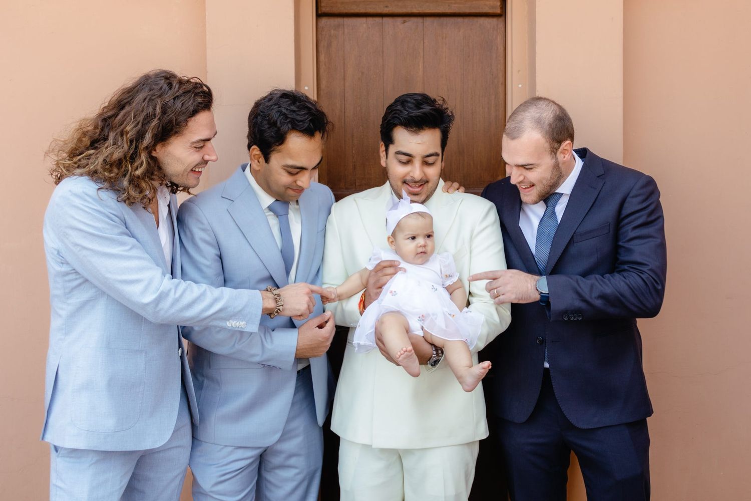 Group of formally dressed people gather around and interact with a baby in white.