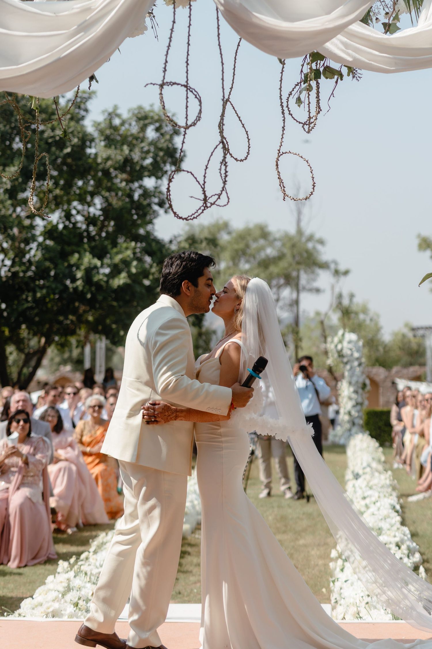 A romantic outdoor wedding ceremony with guests in pink dresses watching the couple share their first kiss.