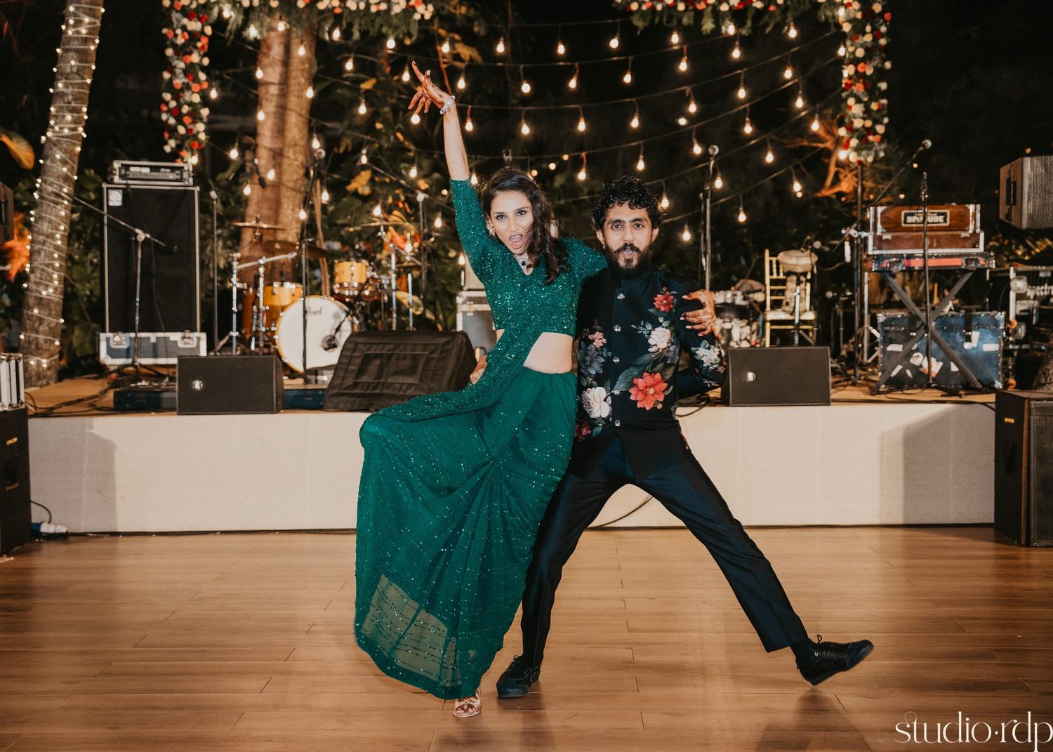 A couple performs a dramatic dance move on a wooden floor with string lights and stage equipment in background.