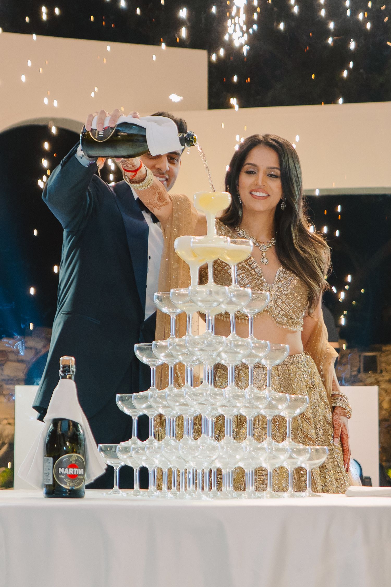 Champagne tower being filled at an elegant event with sparkling lights in the background.