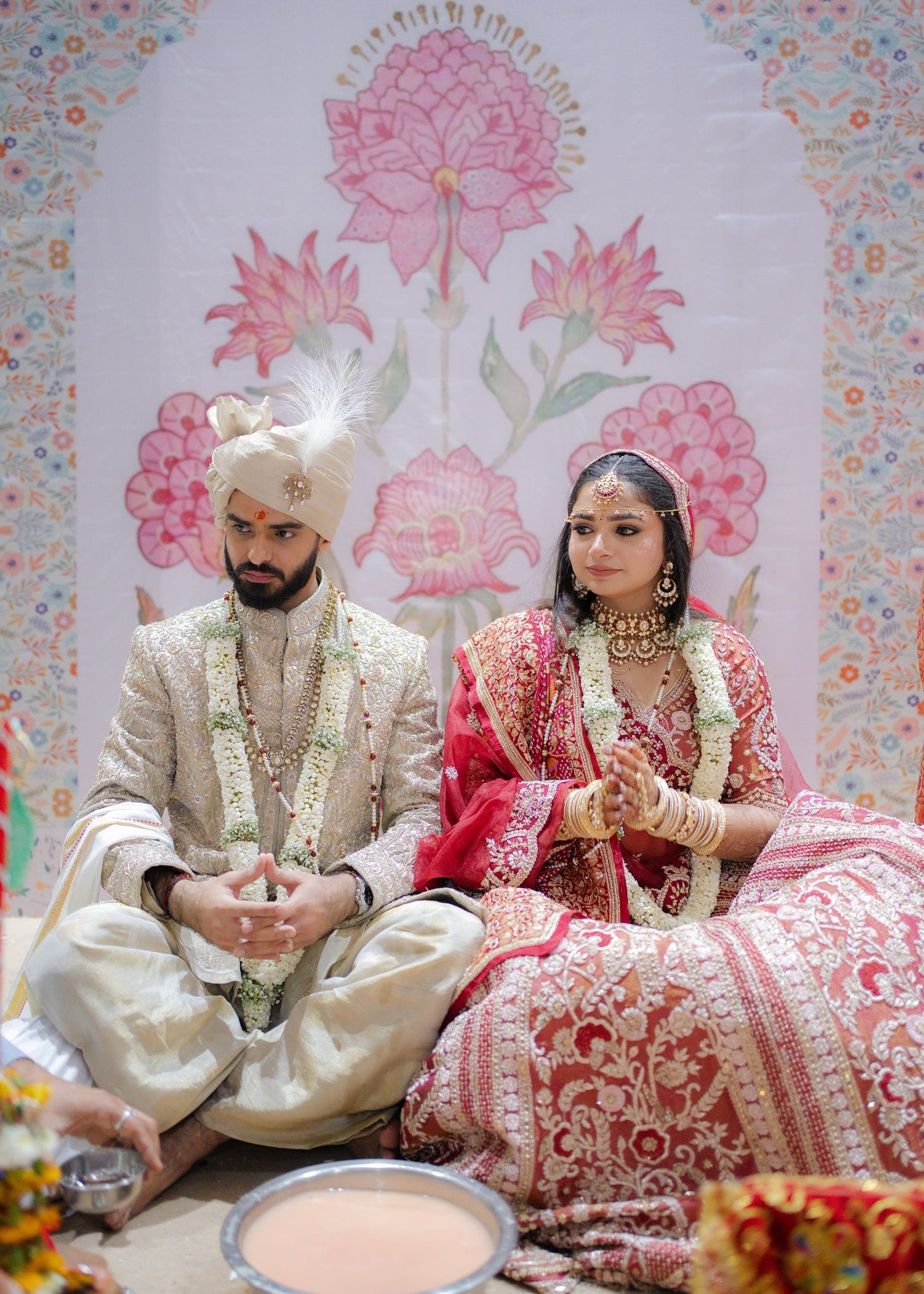 Couple in traditional Indian wedding attire sit together during Hindu marriage ceremony with floral backdrop.