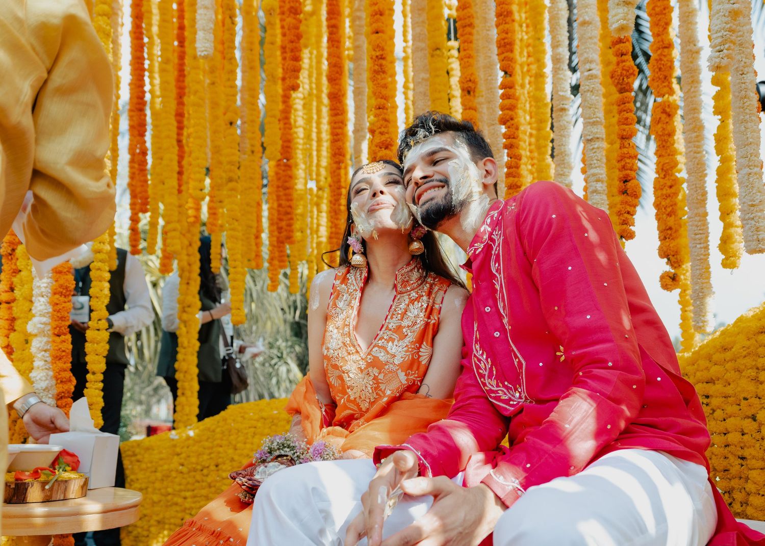 A couple in traditional Indian attire shares a joyful moment under vibrant orange marigold decorations at a celebration.