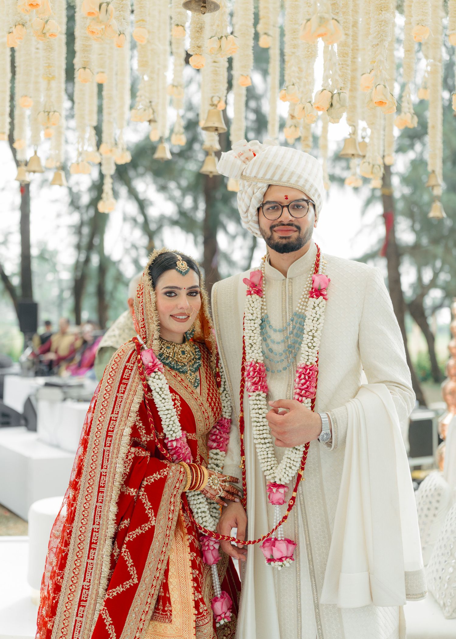 Traditional Indian wedding ceremony with couple in red lehenga and ivory sherwani under hanging floral decor.