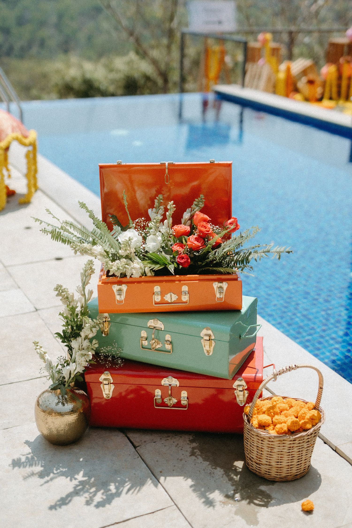 Vintage suitcases stacked by pool with flowers and a wicker basket of oranges on a sunny patio.