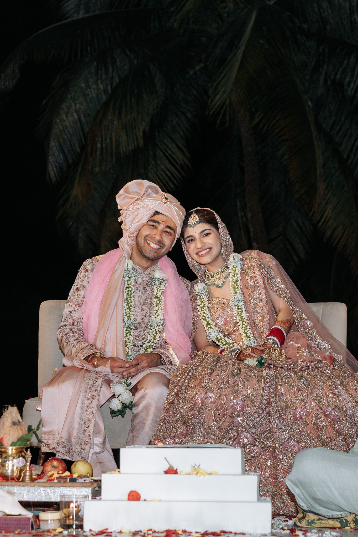 A couple in traditional wedding attire sitting together at their reception table decorated with floral arrangements and candles.