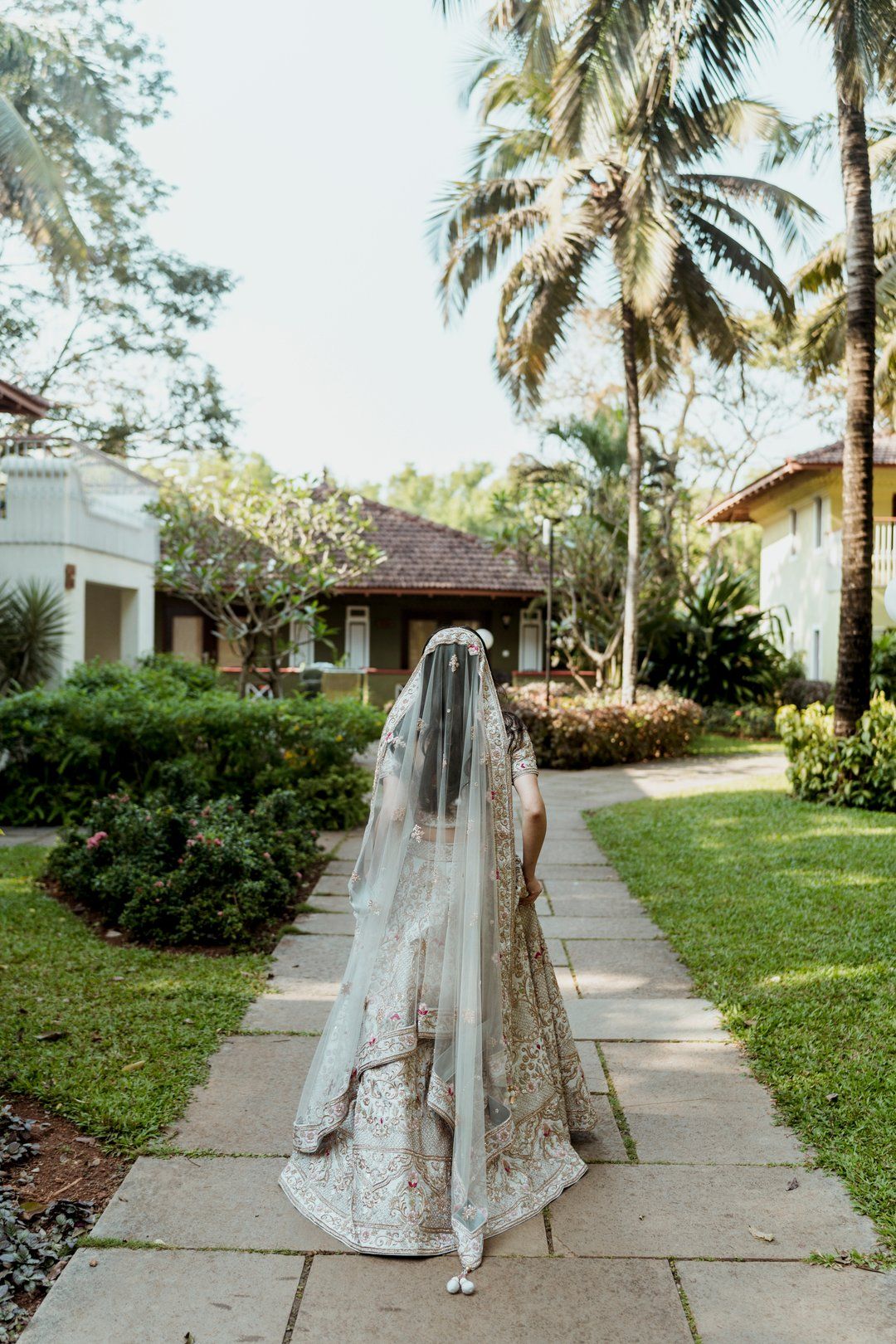 Wedding dress train flowing along a palm-lined pathway in a tropical garden setting.