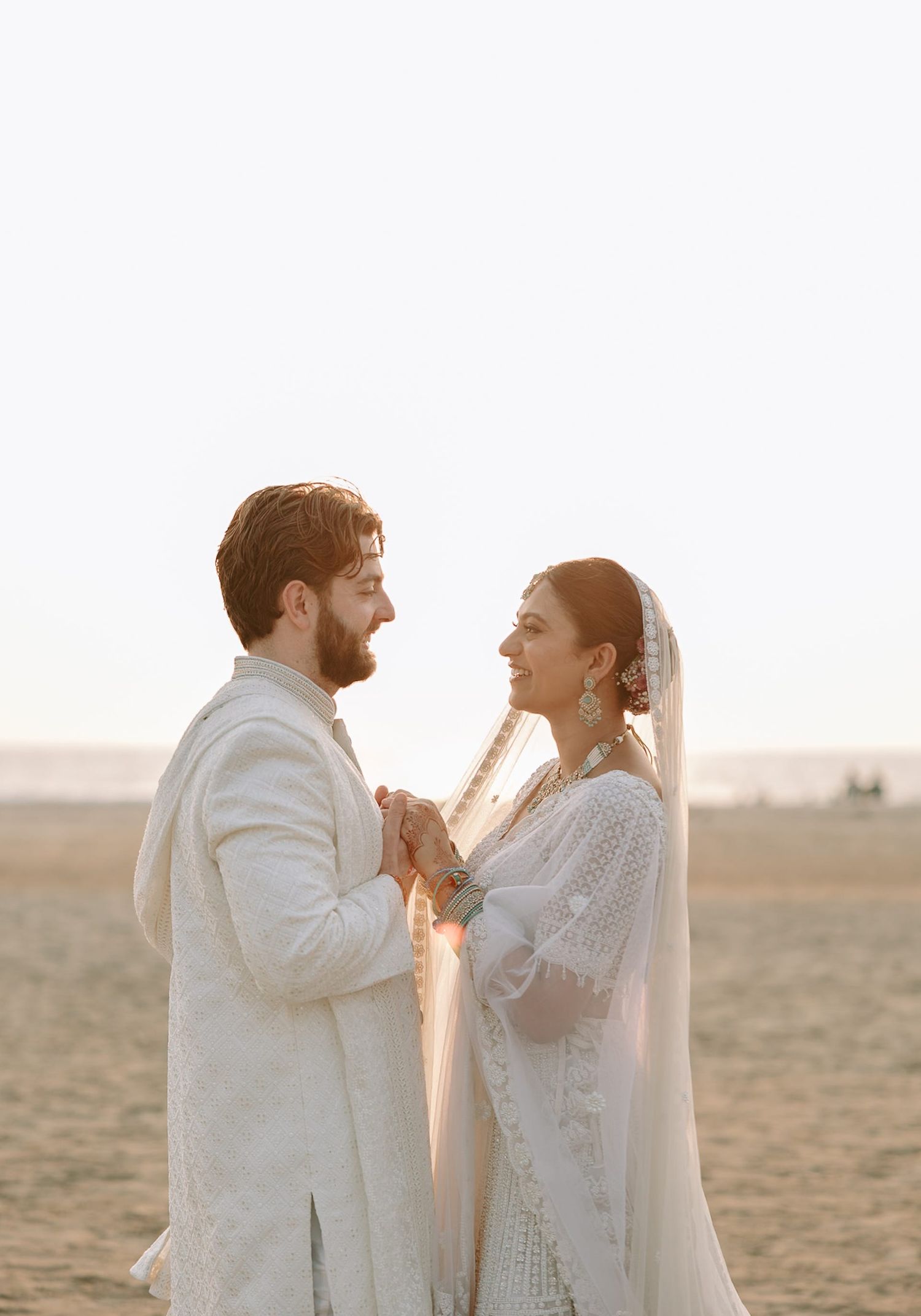 A couple in white South Asian wedding attire holds hands on a sandy beach, smiling at each other in golden hour light.