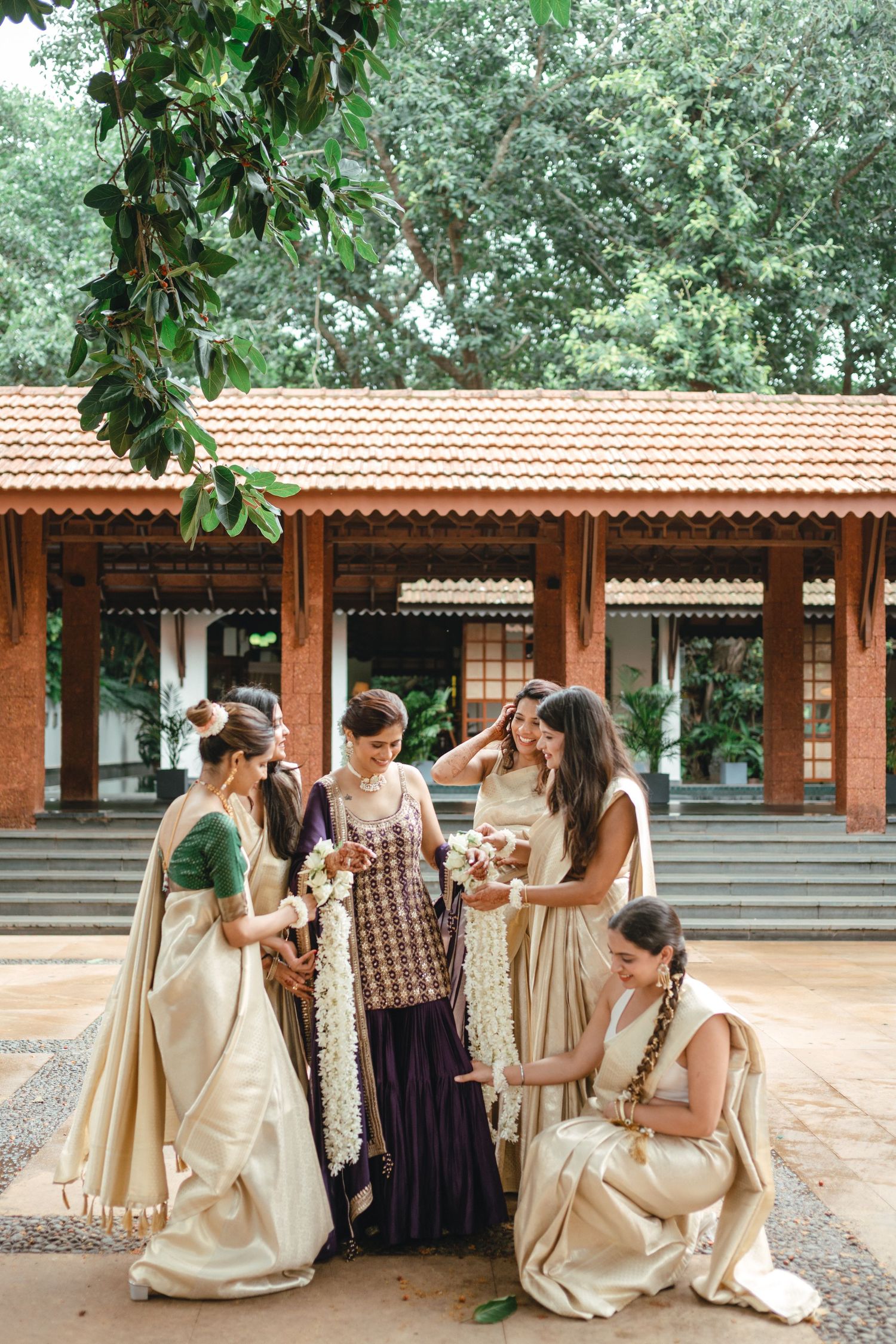Women in cream sarees gather outdoors near a traditional Indian building with red tile roof, surrounded by lush greenery.