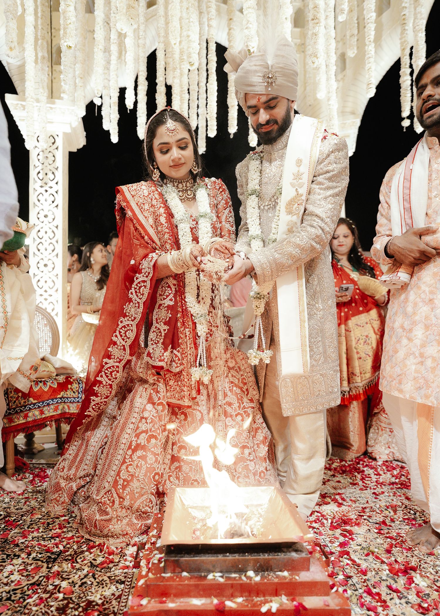Indian bride in red lehenga and groom in cream sherwani performing sacred fire ritual at elegant wedding.