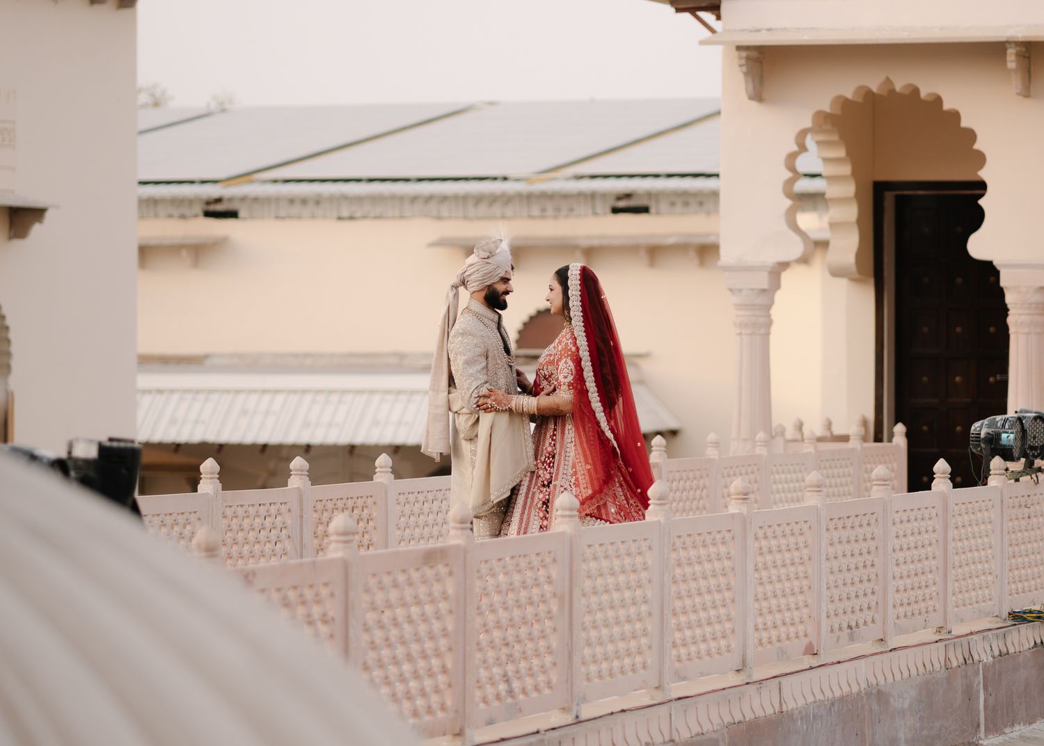 Indian bride and groom in traditional wedding attire share a romantic moment on an ornate white palace terrace.