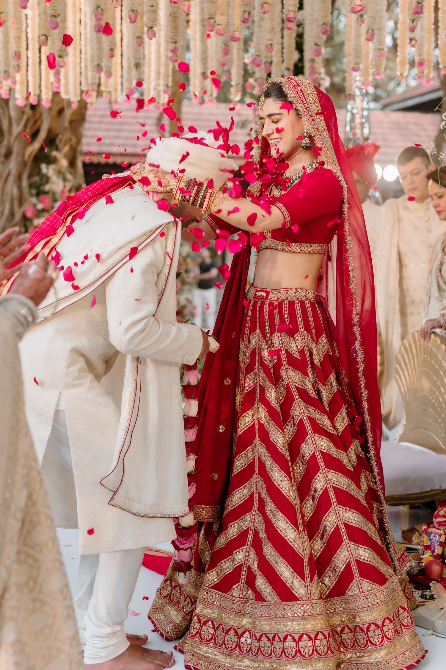 A couple celebrates at a traditional Indian wedding ceremony with red and white decor and rose petal confetti.