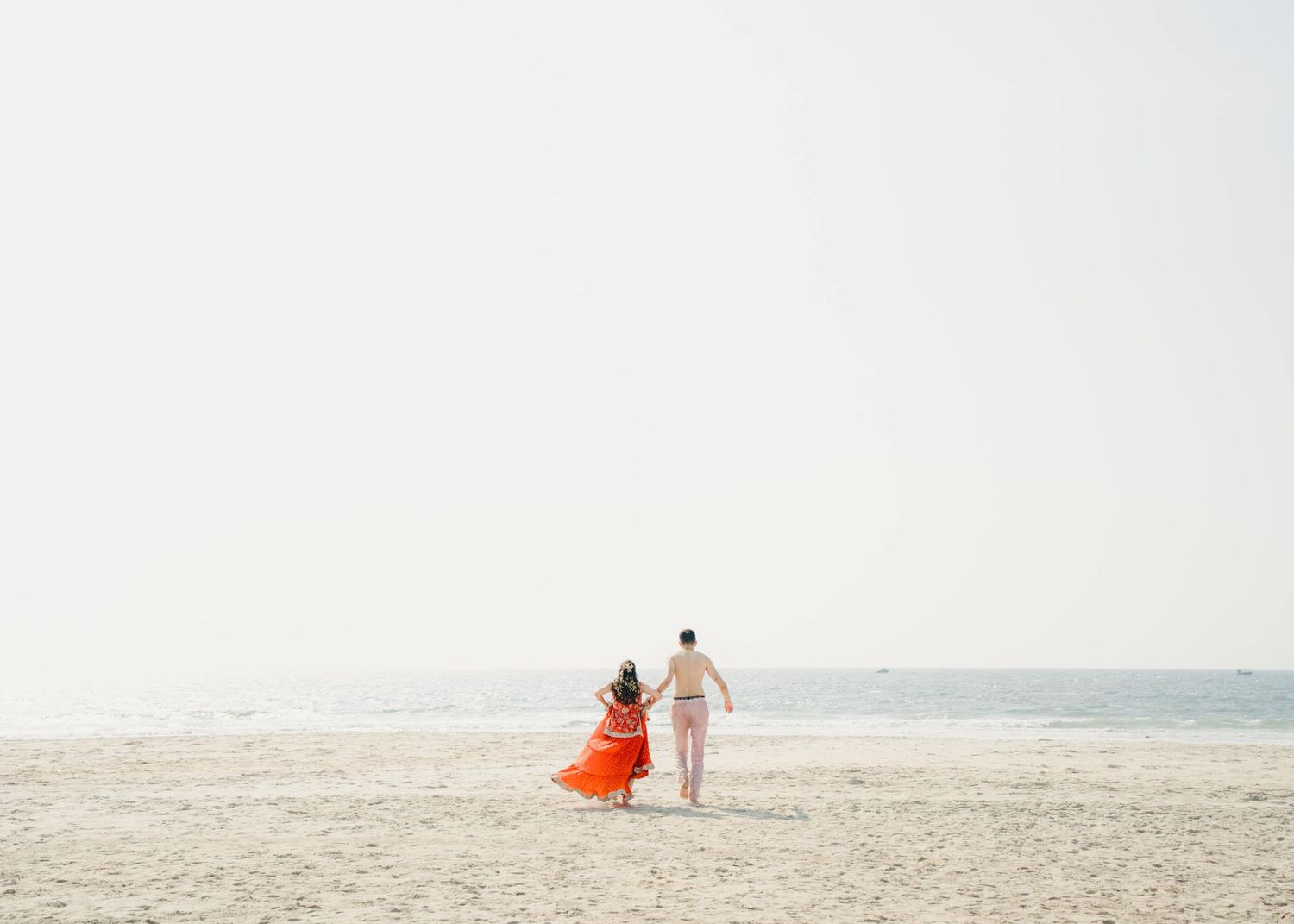 Two figures walk along an empty beach at sunset, one wearing a flowing orange dress and the other in light attire.