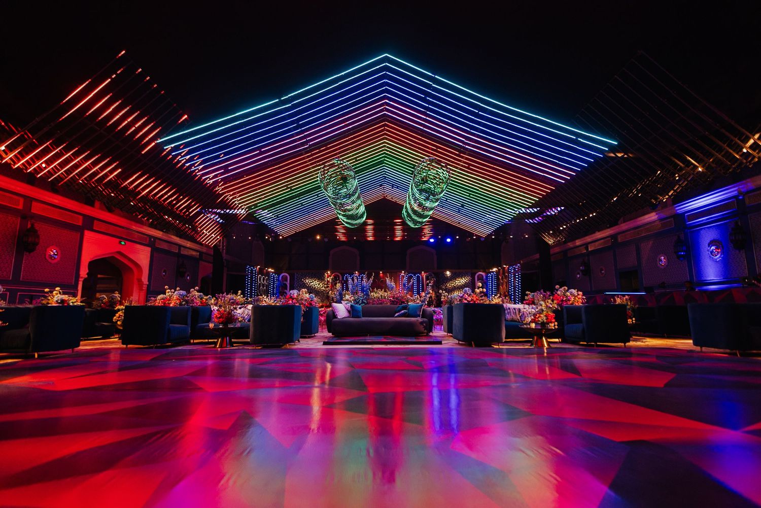 Colorful rainbow neon lights illuminate a nightclub dance floor with red and blue ambient lighting creating a vibrant atmosphere.