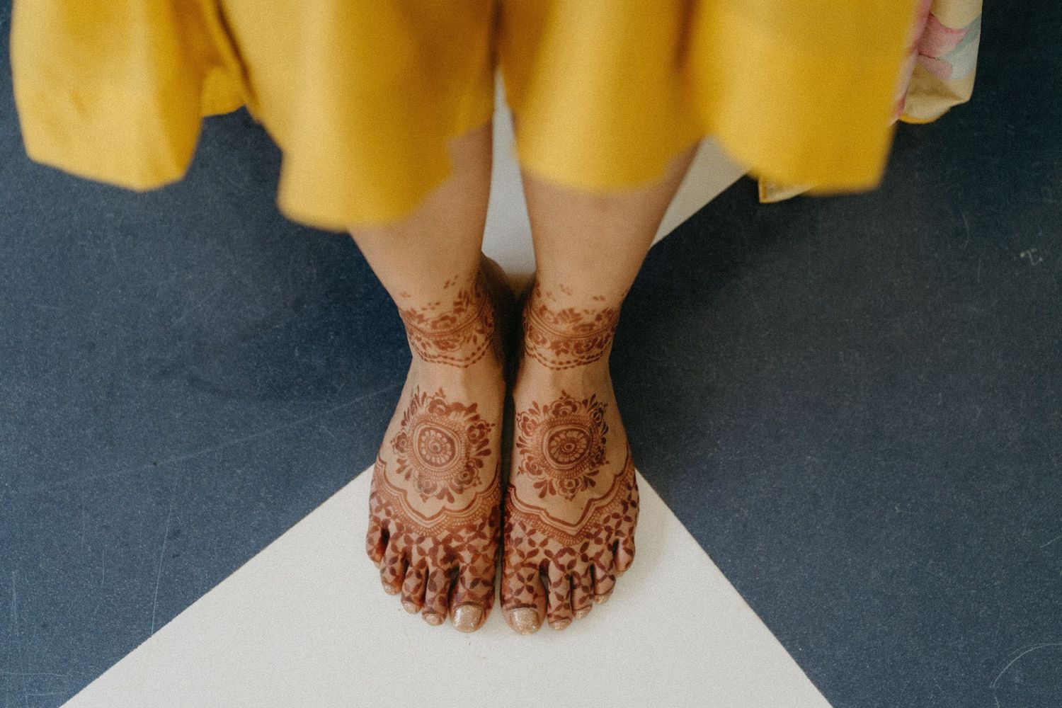 A pair of feet adorned with intricate henna designs rests on a dark floor next to a flowing yellow garment.