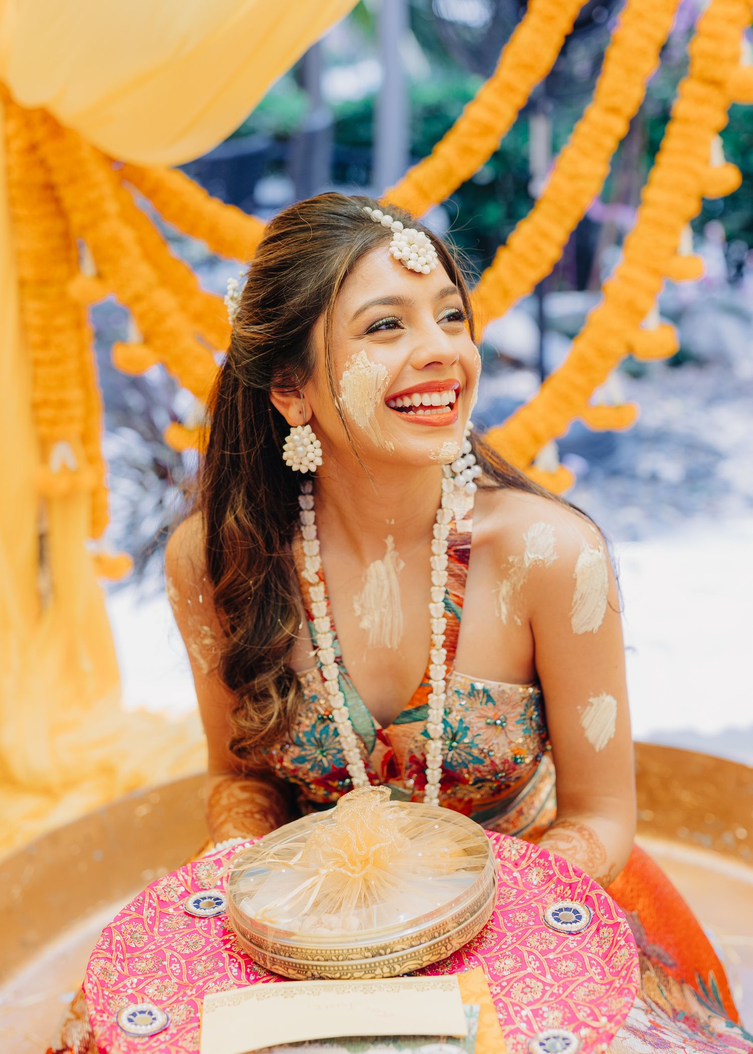 A person wearing traditional Indian attire and jewelry smiles while celebrating during a colorful wedding ceremony.