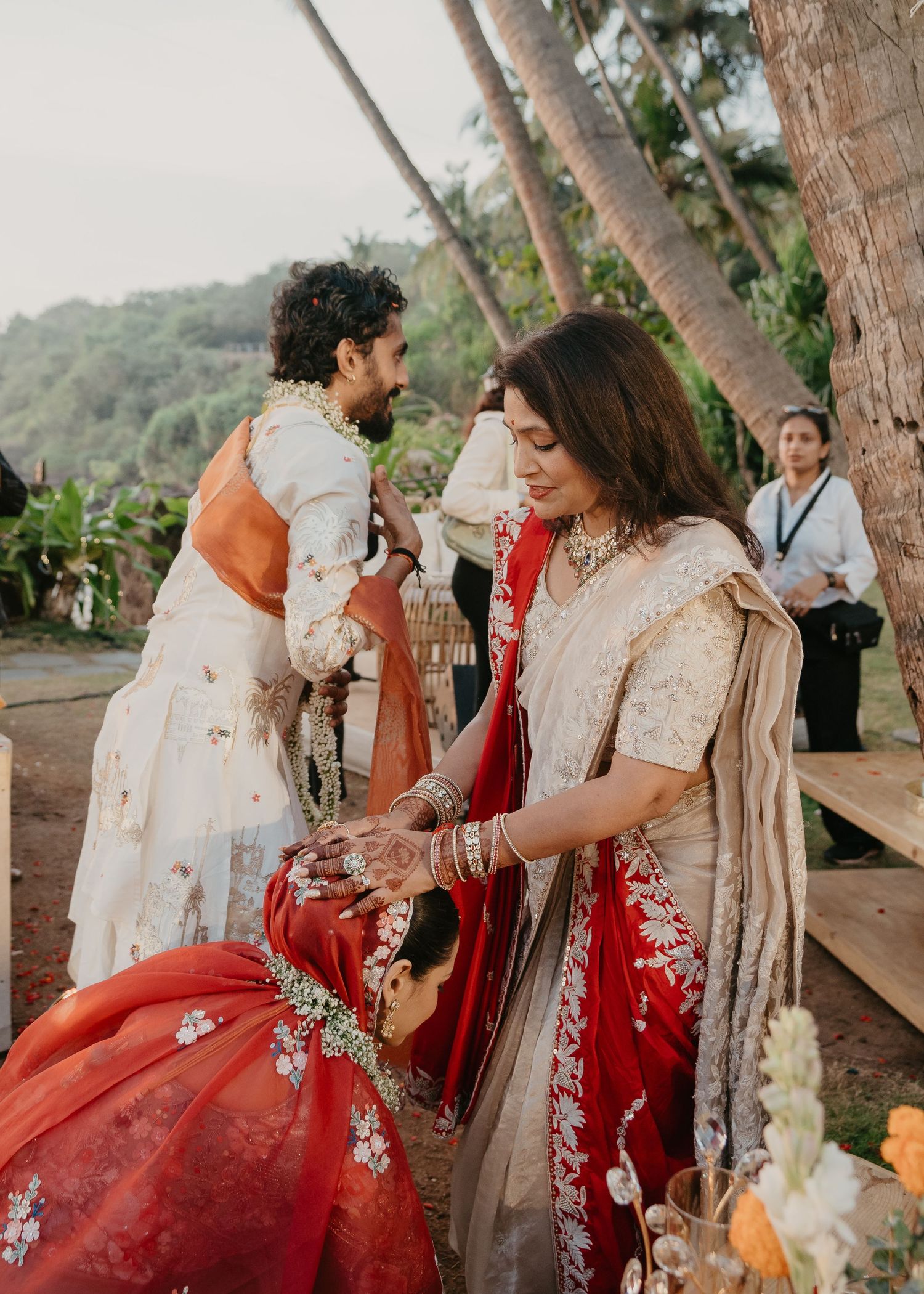 Indian wedding ceremony outdoors with bride in red and gold lehenga, groom in white sherwani, amid tropical palm trees.