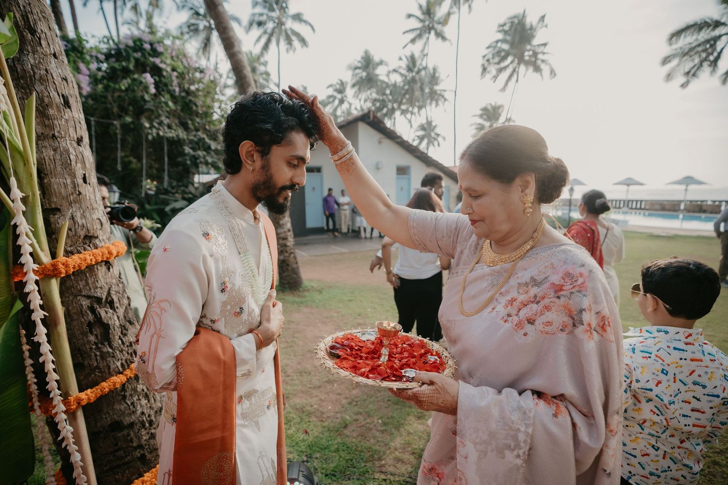 A woman in floral saree applies tilak on a man in traditional kurta at an outdoor Indian wedding ceremony.