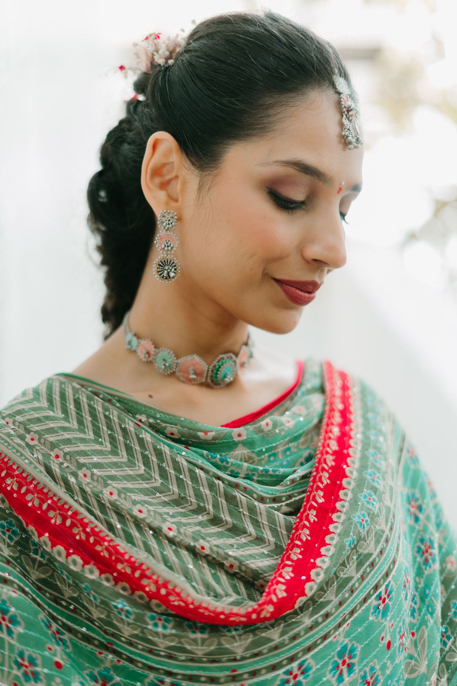 Woman in teal saree with red border, silver jewelry, maang tikka, and floral hair accessories, looking down gracefully.