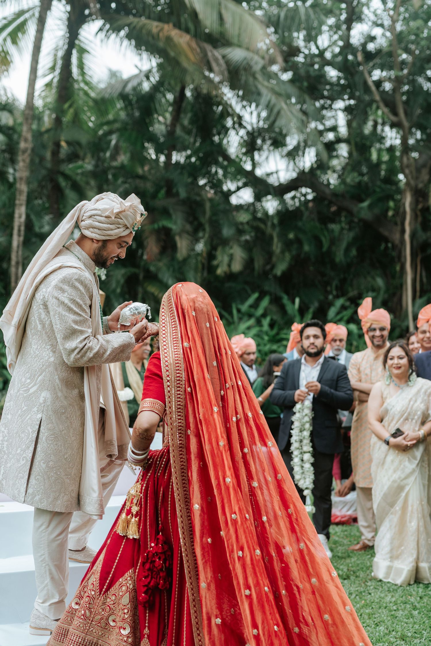 Groom in cream sherwani performs Indian wedding ritual with bride in red lehenga outdoors amid lush tropical greenery.