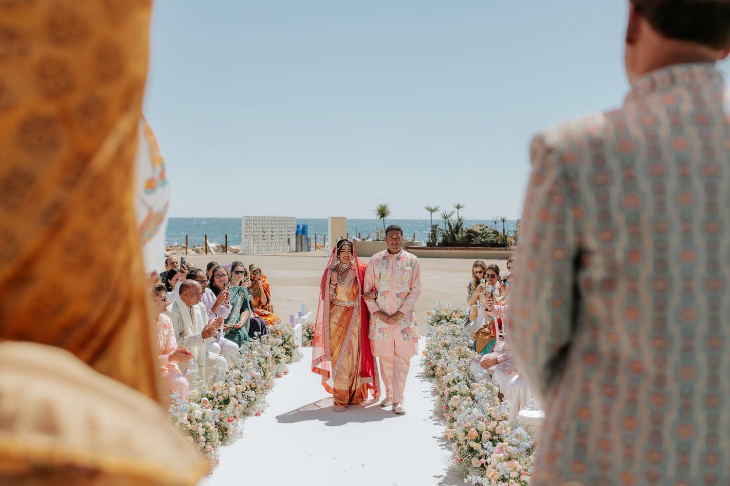 Indian bride and groom walk down floral aisle during outdoor beach Hindu wedding ceremony with guests seated.
