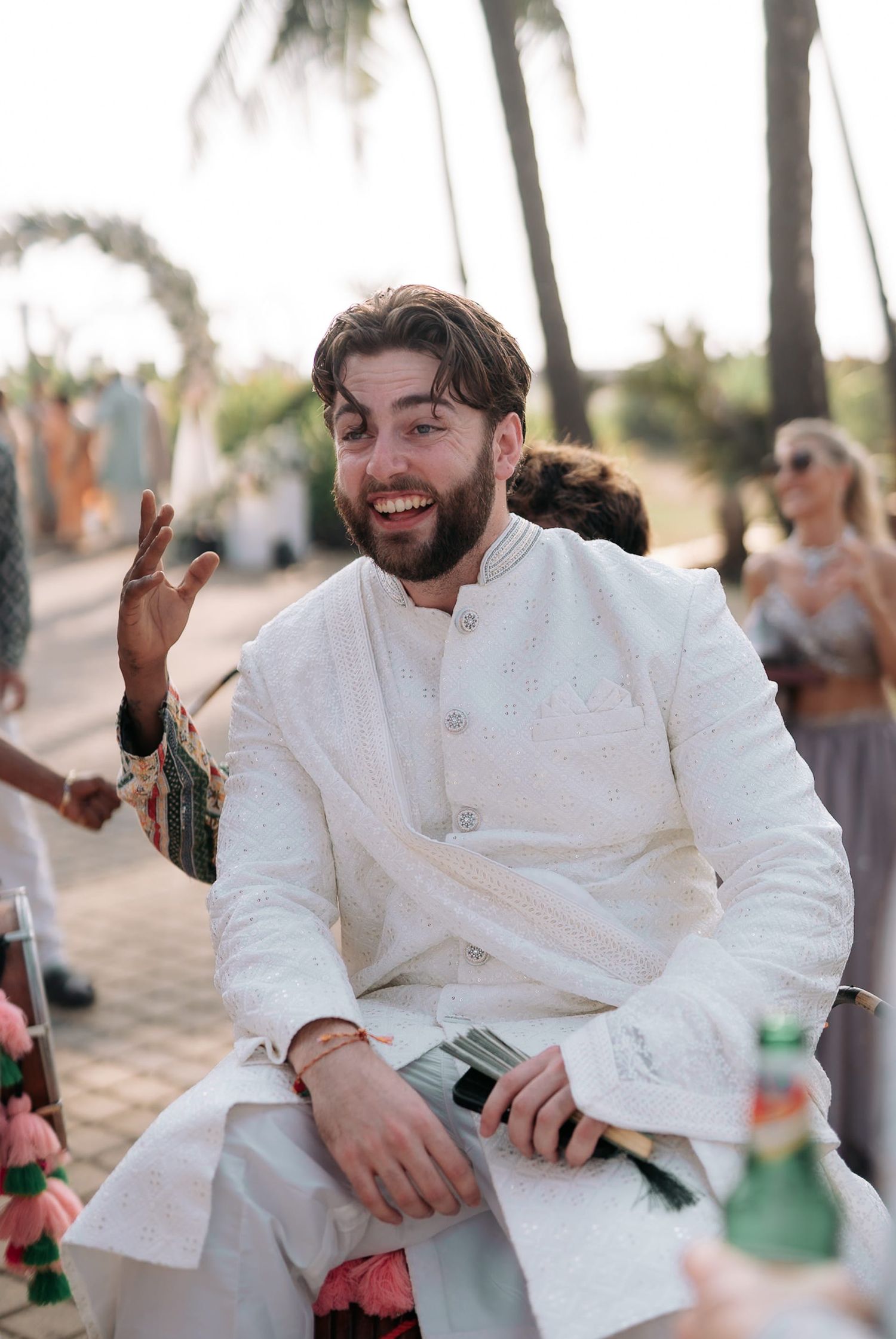 Smiling groom in white sherwani gesturing at an outdoor Indian wedding ceremony with palm trees in background.
