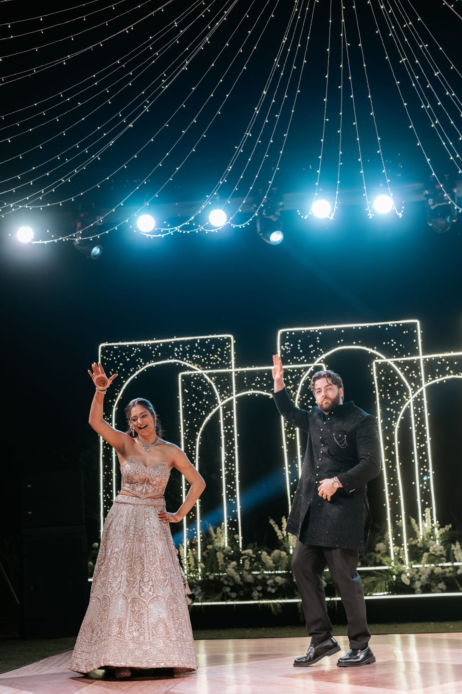 Bride and groom dancing on stage at Indian wedding reception with fairy lights and arch decor backdrop.