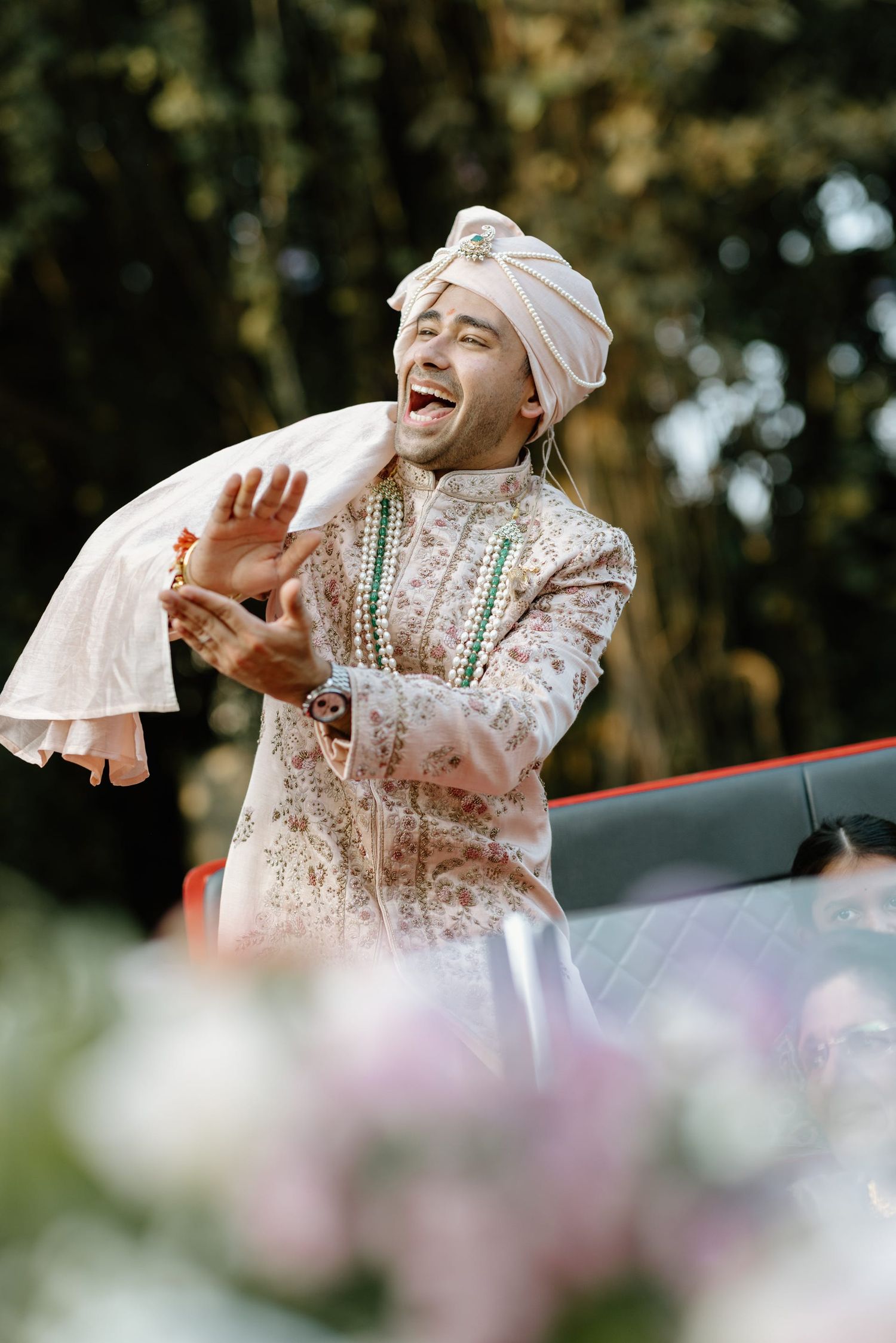 Person in traditional white dress and headscarf laughing expressively outdoors with blurred floral background.