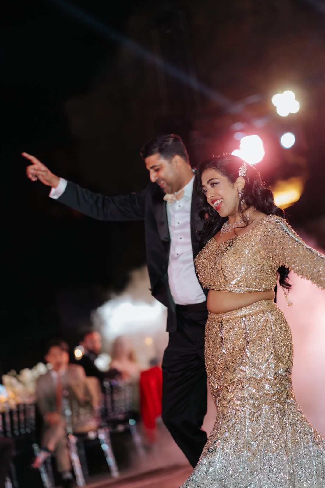 Indian bride and groom making grand entrance on dance floor, bride in gold lehenga under stage lights.