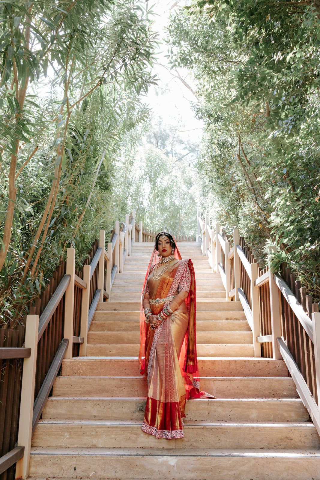 Indian bride in red and gold saree standing on wooden stairs surrounded by lush bamboo trees.