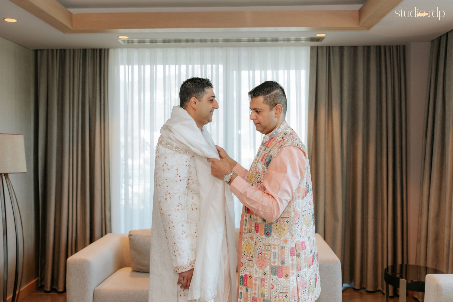 Two grooms in traditional Indian wedding attire help each other dress in an elegant hotel room.