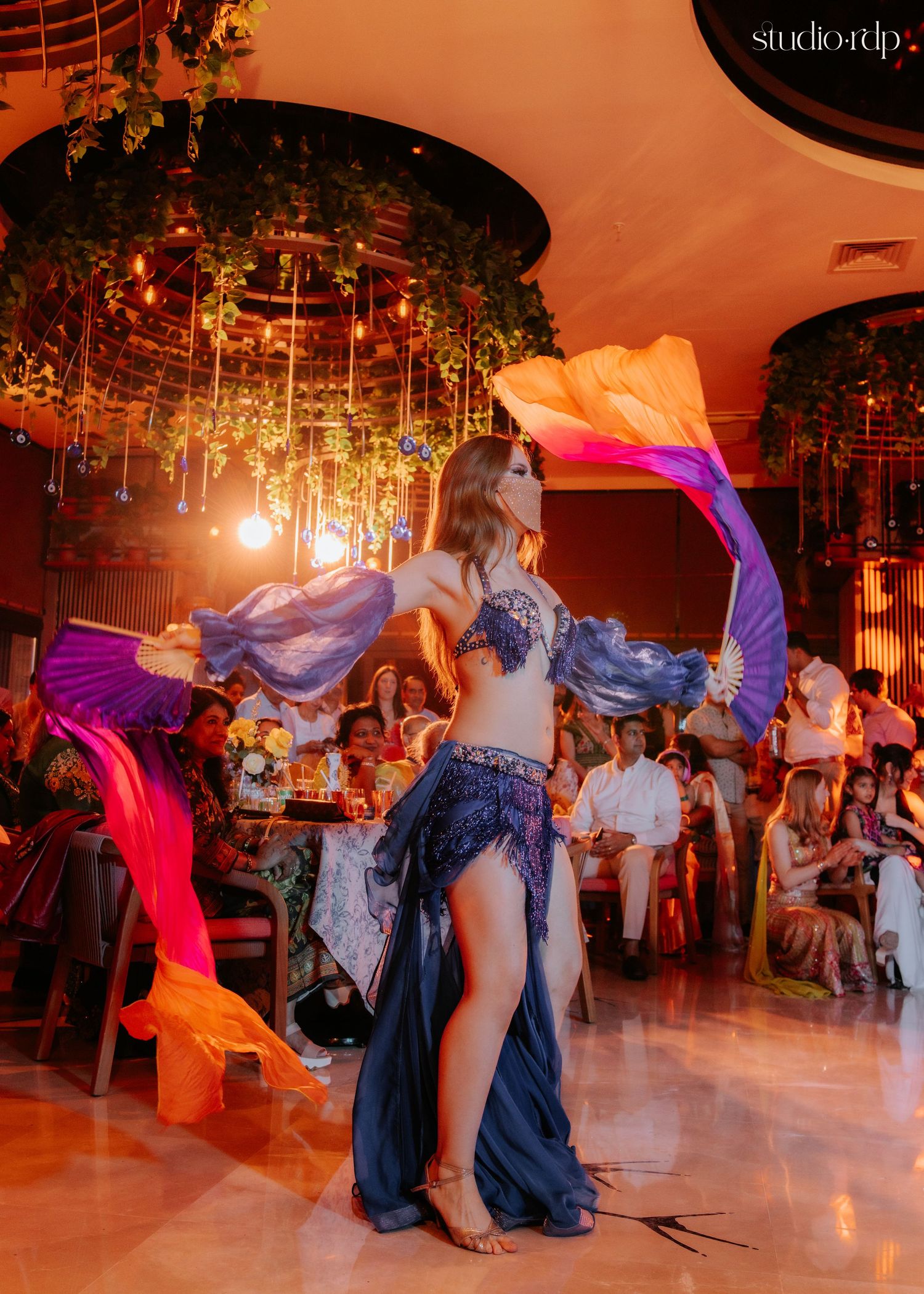 Belly dancer performing with colorful silk fans in a luxurious restaurant setting with lush greenery decor.