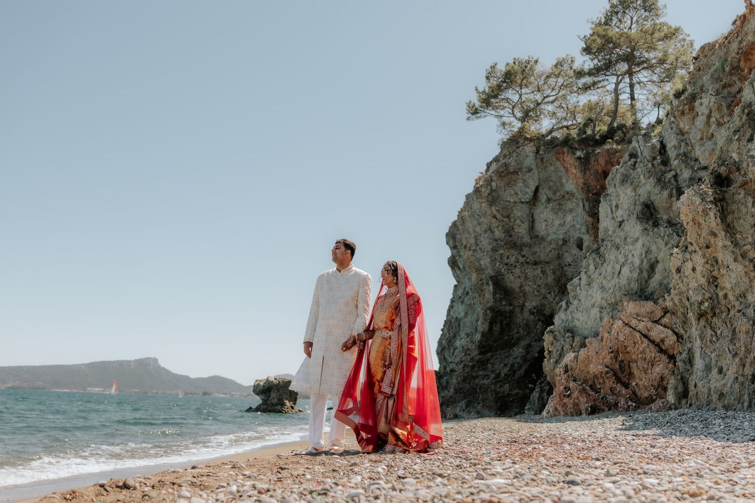 Couple in traditional Indian wedding attire walking hand in hand on a rocky beach with clear blue skies.