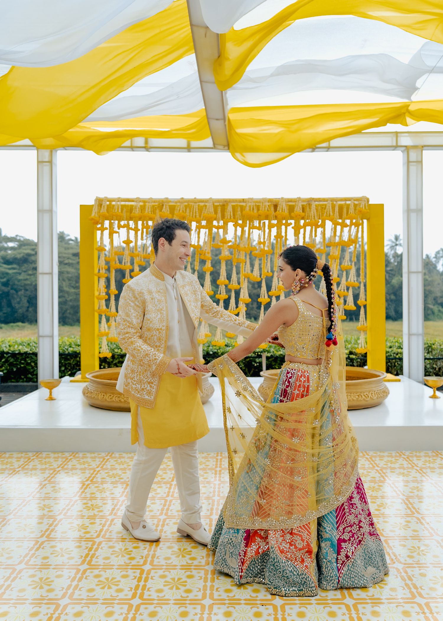 Couple in traditional Indian wedding attire dancing at a vibrant yellow-decorated outdoor ceremony venue.