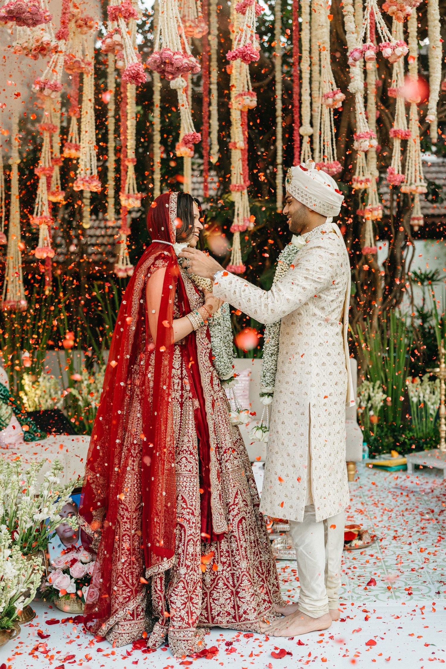 Indian bride in red lehenga and groom in ivory sherwani exchange garlands under floral decorations at traditional wedding.