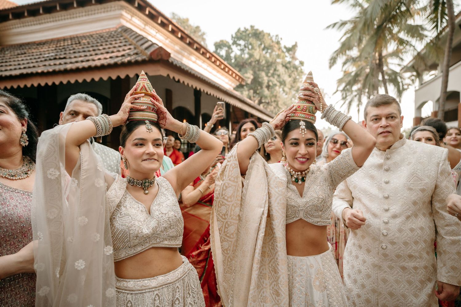 Women in white lehengas dance joyfully during a traditional Indian wedding ceremony outdoors.