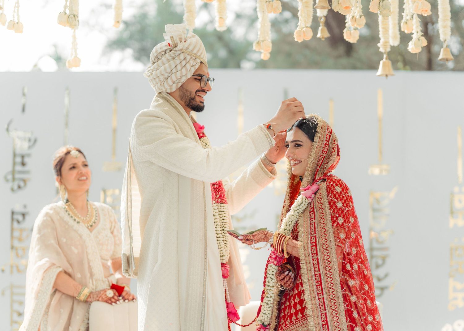 Traditional Indian wedding ceremony with couple in elegant red and ivory attire under hanging floral decorations.