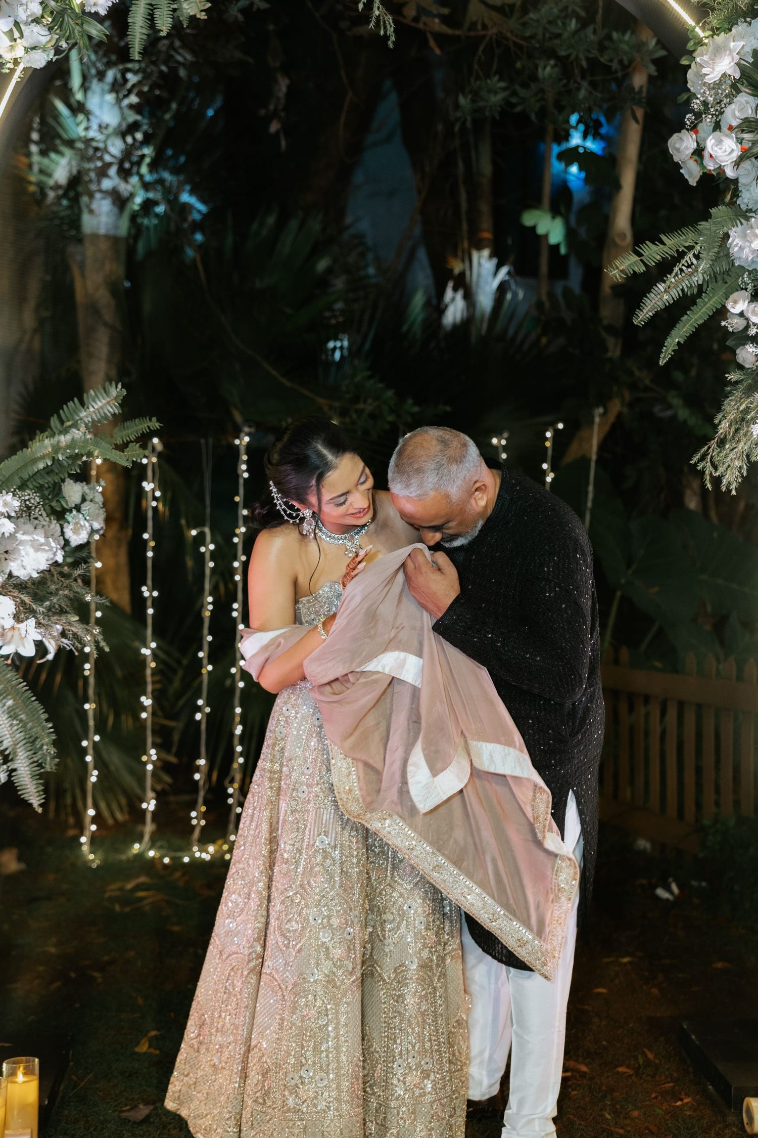Couple embracing under floral arch with fairy lights at outdoor evening wedding ceremony in elegant attire.