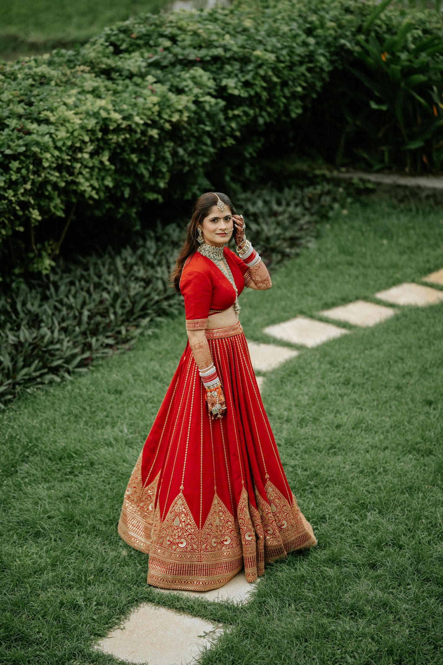 Indian bride in stunning red and gold lehenga with traditional bridal jewelry posing in lush garden setting.