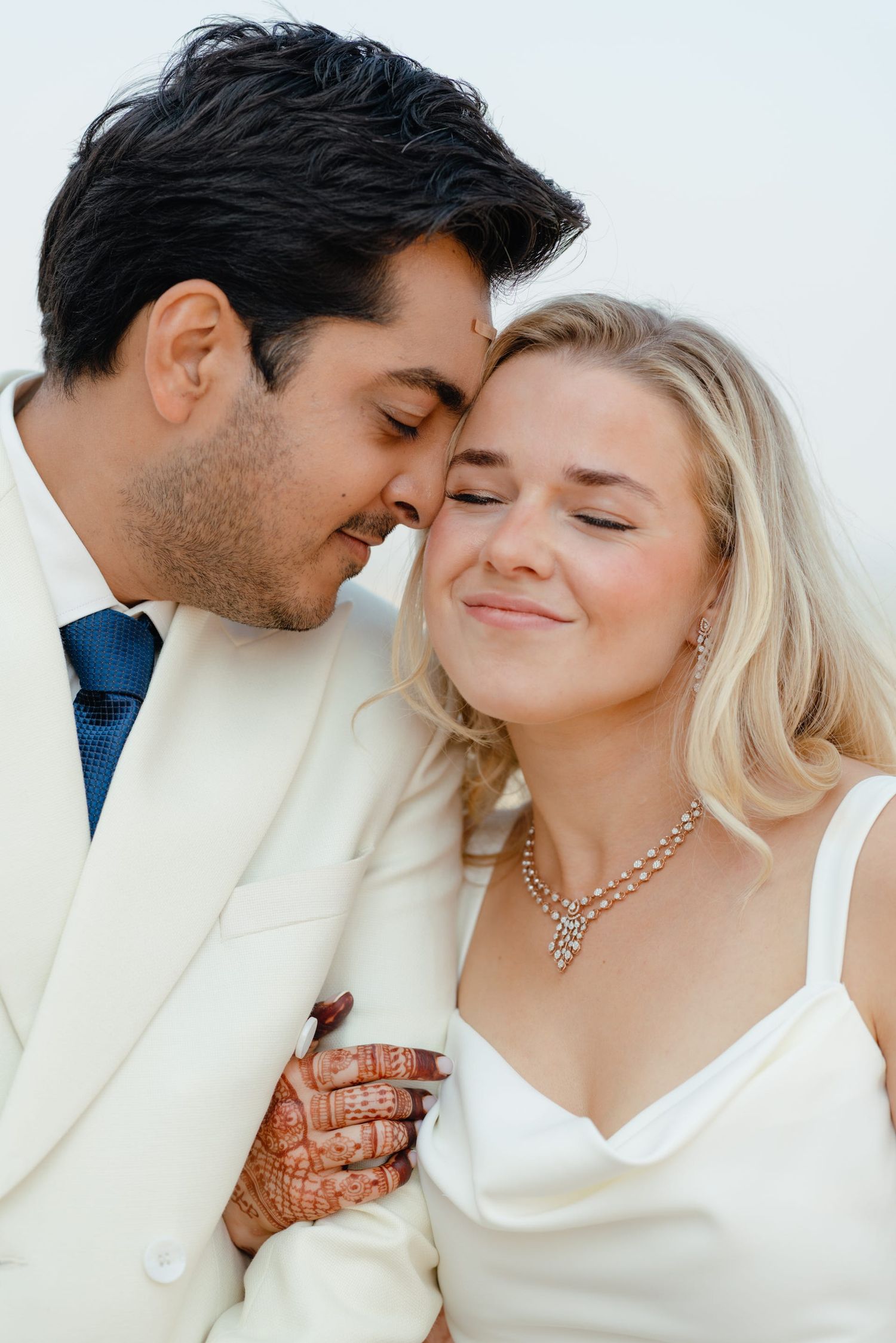 Intimate romantic moment between newlyweds in white attire against bright background.
